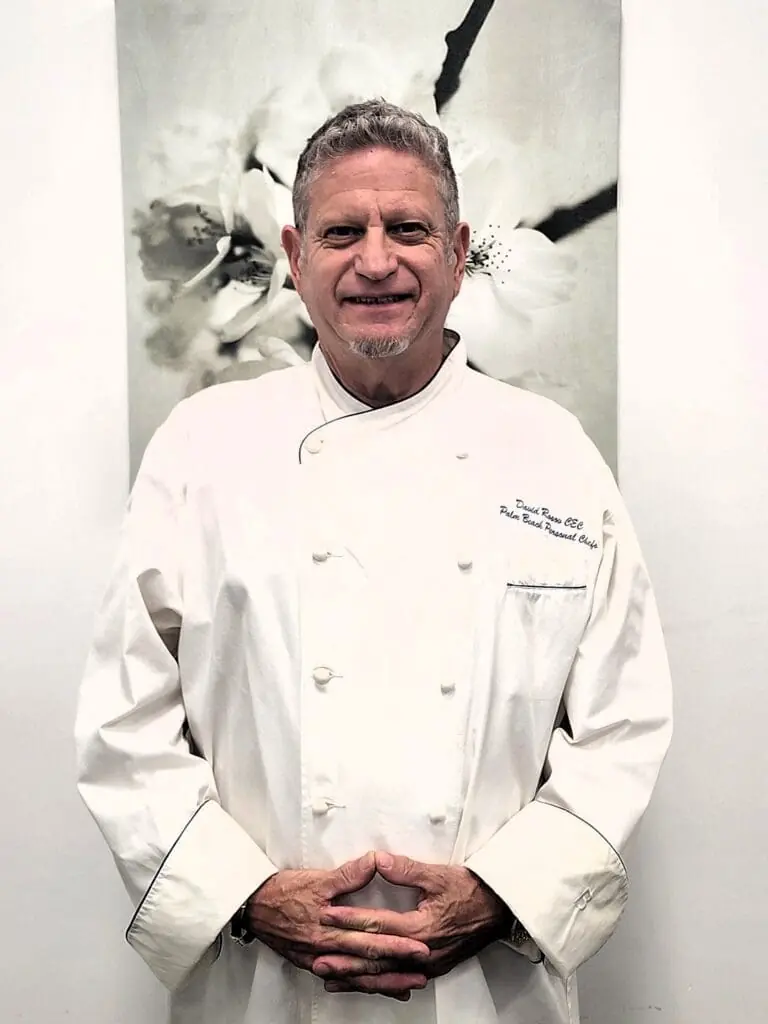 Professional chef in white uniform smiling at camera, offering elite nutrition chef services and personal meal preparation